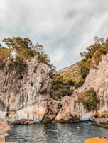 A natural swimming area surrounded by tall rocky cliffs and lush green trees. Several people are enjoying the water, and a small wooden platform is visible near the edge of the pool. The landscape has an earthy, rugged feel with vegetation covering parts of the rocks.