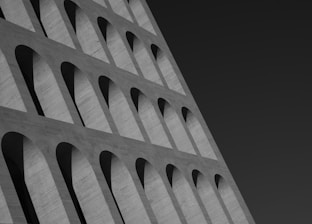 An architectural detail of a modern building featuring a series of repetitive arch patterns arranged in a slanted perspective. The structures are composed of concrete, creating a stark contrast with the dark sky in the background.