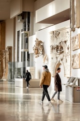 A museum gallery with several people walking and observing. The walls are adorned with classical sculptures and reliefs. The space is well-lit, with large glass panels and light-colored interiors. The floor is polished and reflective.