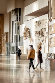 A museum gallery with several people walking and observing. The walls are adorned with classical sculptures and reliefs. The space is well-lit, with large glass panels and light-colored interiors. The floor is polished and reflective.