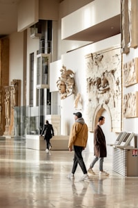 A museum gallery with several people walking and observing. The walls are adorned with classical sculptures and reliefs. The space is well-lit, with large glass panels and light-colored interiors. The floor is polished and reflective.