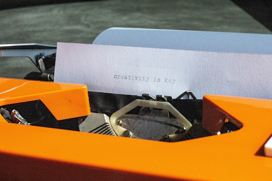 A close-up of a typewriter with a piece of paper inserted, featuring the words 'creativity is key' typed on it. The typewriter is prominently orange, with visible mechanical parts.