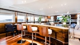 A luxurious yacht interior featuring a sleek bar area with four modern wooden stools, glossy wooden surfaces, and large windows providing ample natural light. The space is decorated with potted green plants and plush seating areas, creating an elegant and inviting atmosphere.