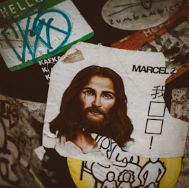 A collage of various stickers and posters is layered on a surface. The most prominent element is a torn, artistic depiction of a face with long hair and a tear on the cheek. Surrounding this image are various stickers, including one with stylized text and a partial cartoon-like drawing in black and white. The arrangement is eclectic and chaotic.
