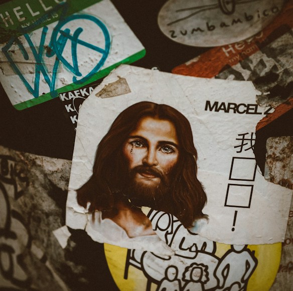 A collage of various stickers and posters is layered on a surface. The most prominent element is a torn, artistic depiction of a face with long hair and a tear on the cheek. Surrounding this image are various stickers, including one with stylized text and a partial cartoon-like drawing in black and white. The arrangement is eclectic and chaotic.