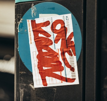 A close-up of a postal label with red graffiti text placed on a black surface with a blue circular background. The label has visible printed text and barcode.