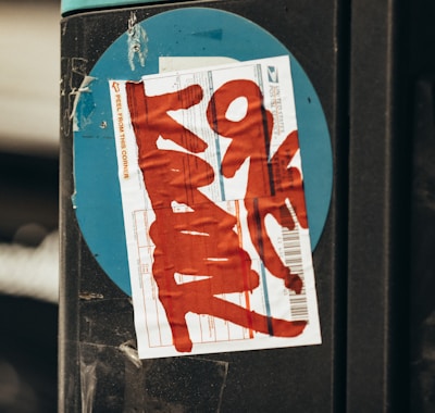 A close-up of a postal label with red graffiti text placed on a black surface with a blue circular background. The label has visible printed text and barcode.