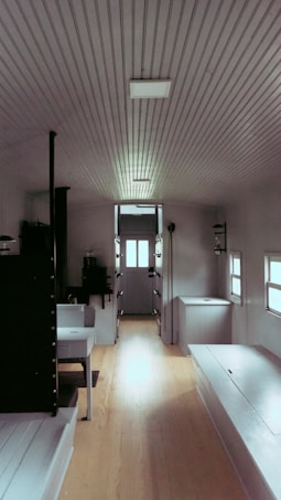 A narrow interior space featuring wooden floors and walls with a few small furniture pieces. The ceiling has a corrugated design with a couple of lights. There are small windows on both sides allowing some natural light.