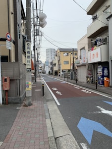 A narrow urban street flanked by low-rise buildings, with overhead power lines and signs in a foreign language. The street is empty, with bicycles parked on the side, some laundry hanging on a balcony, and a red mailbox visible. A larger apartment building is seen in the background.