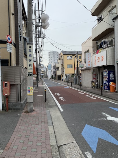 A narrow urban street flanked by low-rise buildings, with overhead power lines and signs in a foreign language. The street is empty, with bicycles parked on the side, some laundry hanging on a balcony, and a red mailbox visible. A larger apartment building is seen in the background.