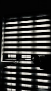 Horizontal blinds partially cover a window, creating a pattern of alternating light and shadow. Below the window, there is a reflective surface, possibly a table, displaying the same pattern along with a few small objects.