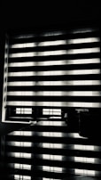 Horizontal blinds partially cover a window, creating a pattern of alternating light and shadow. Below the window, there is a reflective surface, possibly a table, displaying the same pattern along with a few small objects.