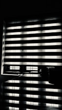 Horizontal blinds partially cover a window, creating a pattern of alternating light and shadow. Below the window, there is a reflective surface, possibly a table, displaying the same pattern along with a few small objects.