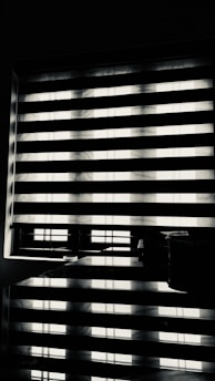 Horizontal blinds partially cover a window, creating a pattern of alternating light and shadow. Below the window, there is a reflective surface, possibly a table, displaying the same pattern along with a few small objects.