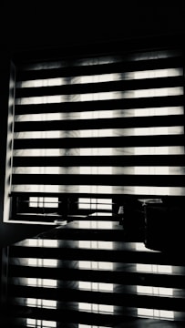 Horizontal blinds partially cover a window, creating a pattern of alternating light and shadow. Below the window, there is a reflective surface, possibly a table, displaying the same pattern along with a few small objects.