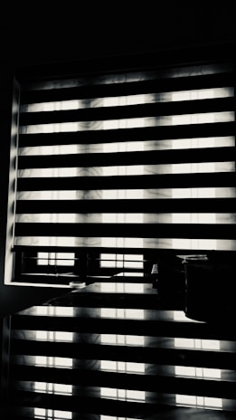Horizontal blinds partially cover a window, creating a pattern of alternating light and shadow. Below the window, there is a reflective surface, possibly a table, displaying the same pattern along with a few small objects.