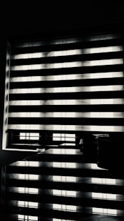 Horizontal blinds partially cover a window, creating a pattern of alternating light and shadow. Below the window, there is a reflective surface, possibly a table, displaying the same pattern along with a few small objects.