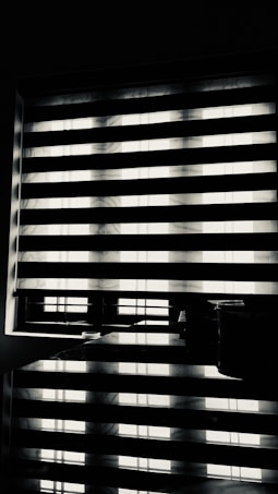 Horizontal blinds partially cover a window, creating a pattern of alternating light and shadow. Below the window, there is a reflective surface, possibly a table, displaying the same pattern along with a few small objects.