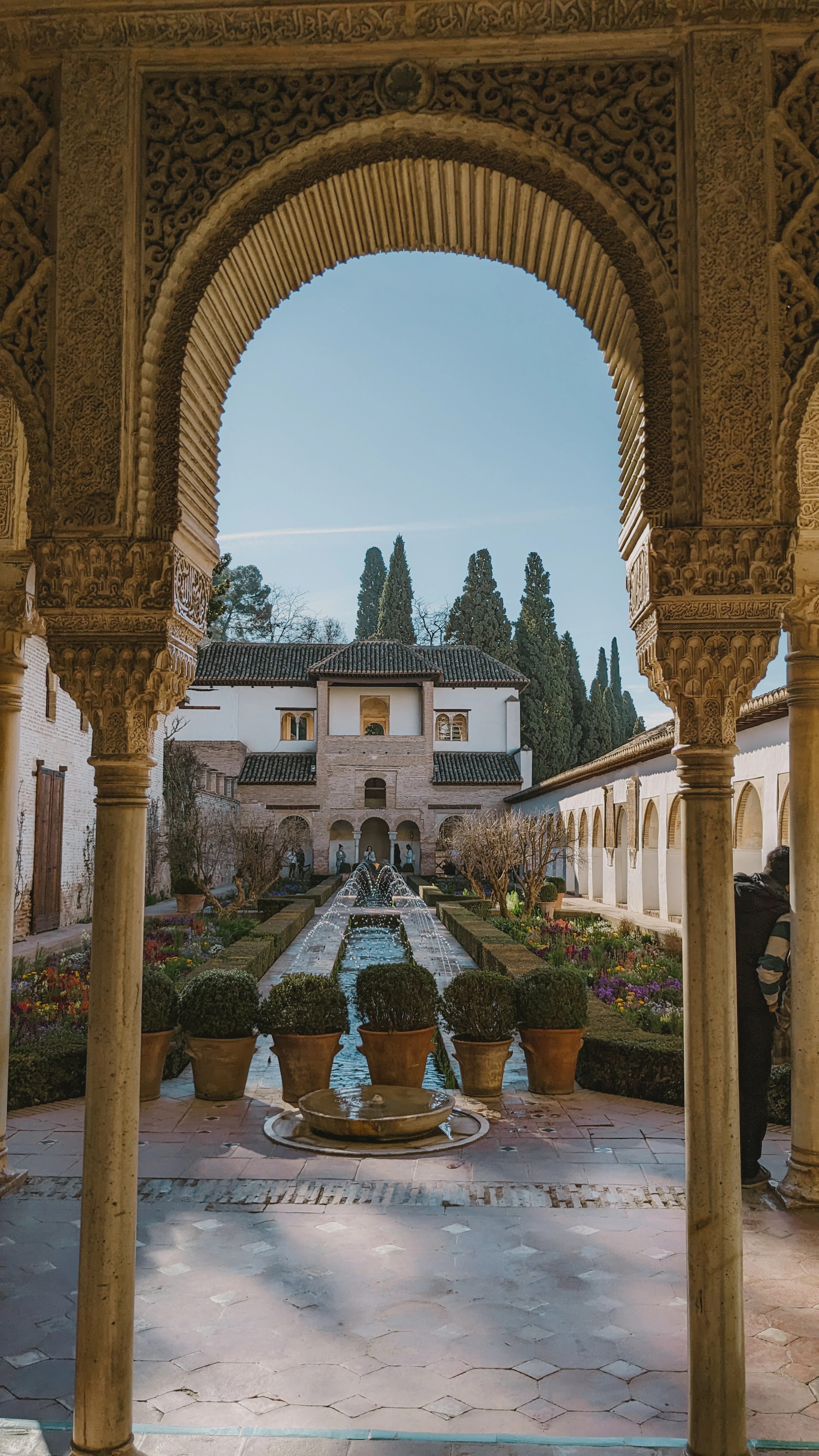 A carved Generalife arch frames a garden courtyard with clipped hedges, potted plants and a long reflecting fountain.