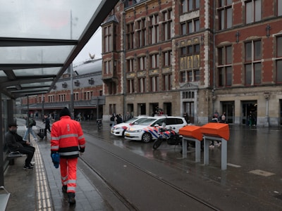 A person in bright orange work attire walks along a sidewalk near a bus stop with a modern glass canopy. Several people can be seen sitting or standing nearby. In the background, a historic brick building with elaborate architectural details is visible. There are vehicles, including a police car and a motorcycle, parked near the building. The scene is overcast and the ground appears wet from rain.