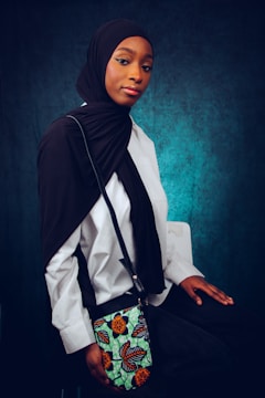A woman is sitting in a studio setting, wearing a black headscarf and a white blouse. She has a colorful patterned shoulder bag and the background is a textured teal color.