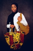 A person wearing a white shirt and a black headscarf is standing against a dark backdrop, holding two vibrant, patterned bags. The bags feature bold, colorful designs with yellow floral and abstract patterns on a red and black background.