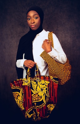A person wearing a white shirt and a black headscarf is standing against a dark backdrop, holding two vibrant, patterned bags. The bags feature bold, colorful designs with yellow floral and abstract patterns on a red and black background.