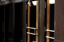 A close-up view of several gold-colored metal doors with vertical handles, set within a dark frame. The doors have reflective glass panels that are partially shaded, suggesting a quiet or closed-off environment. The lighting emphasizes the metallic sheen of the door handles and frames.