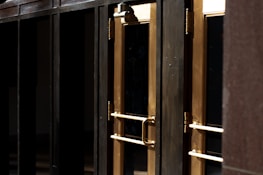 A close-up view of several gold-colored metal doors with vertical handles, set within a dark frame. The doors have reflective glass panels that are partially shaded, suggesting a quiet or closed-off environment. The lighting emphasizes the metallic sheen of the door handles and frames.