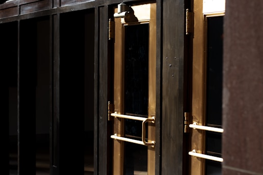 A close-up view of several gold-colored metal doors with vertical handles, set within a dark frame. The doors have reflective glass panels that are partially shaded, suggesting a quiet or closed-off environment. The lighting emphasizes the metallic sheen of the door handles and frames.
