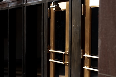 A close-up view of several gold-colored metal doors with vertical handles, set within a dark frame. The doors have reflective glass panels that are partially shaded, suggesting a quiet or closed-off environment. The lighting emphasizes the metallic sheen of the door handles and frames.