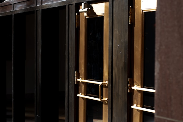 A close-up view of several gold-colored metal doors with vertical handles, set within a dark frame. The doors have reflective glass panels that are partially shaded, suggesting a quiet or closed-off environment. The lighting emphasizes the metallic sheen of the door handles and frames.