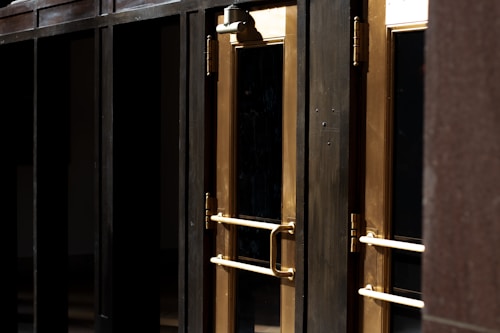 A close-up view of several gold-colored metal doors with vertical handles, set within a dark frame. The doors have reflective glass panels that are partially shaded, suggesting a quiet or closed-off environment. The lighting emphasizes the metallic sheen of the door handles and frames.