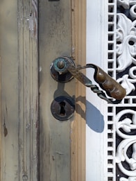 An ornate, vintage door handle with a distressed, peeling wooden door surface is accompanied by intricate white wrought iron work. The handle is made of aged brass with a textured grip and an antique keyhole below it.