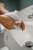 A pair of hands holding a white towel over a bathroom sink with a silver faucet. The sink is white and clean, with a drain visible.