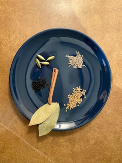 A variety of spices are displayed on a dark blue plate. The plate contains cardamom pods, black cumin seeds, a cinnamon stick, cumin seeds, fenugreek seeds, and bay leaves. The arrangement is neat, with each spice sectioned away from the others for clear visibility. The background features a warm brown surface that contrasts with the cool tone of the plate.