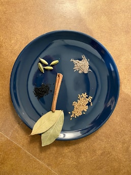 A variety of spices are displayed on a dark blue plate. The plate contains cardamom pods, black cumin seeds, a cinnamon stick, cumin seeds, fenugreek seeds, and bay leaves. The arrangement is neat, with each spice sectioned away from the others for clear visibility. The background features a warm brown surface that contrasts with the cool tone of the plate.