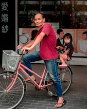An adult wearing a red shirt and jeans is riding a pink bicycle with a child seated in a rear child seat. The child is dressed in a black t-shirt and pink shoes. The bicycle has a light gray basket at the front and is parked on a city street with a building in the background featuring posters of couples in wedding attire.