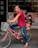 An adult wearing a red shirt and jeans is riding a pink bicycle with a child seated in a rear child seat. The child is dressed in a black t-shirt and pink shoes. The bicycle has a light gray basket at the front and is parked on a city street with a building in the background featuring posters of couples in wedding attire.