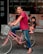 An adult wearing a red shirt and jeans is riding a pink bicycle with a child seated in a rear child seat. The child is dressed in a black t-shirt and pink shoes. The bicycle has a light gray basket at the front and is parked on a city street with a building in the background featuring posters of couples in wedding attire.