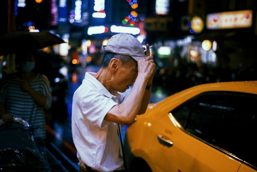 An older man is seen adjusting his white cap while standing beside a yellow car. He is wearing a white shirt and appears to be in a busy urban environment at night, with bright lights and signs in the background. Another person holding an umbrella stands nearby, partially obscured.