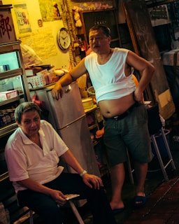Two men are in a cluttered room with warm lighting. One is seated, wearing a white polo shirt, and holding a piece of paper. The other is standing with a hand on his hip, dressed in a white sleeveless shirt and shorts. Behind them is a refrigerator, and various household items are stacked on shelves.
