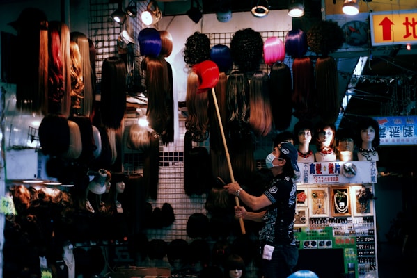 A person wearing a mask is standing indoors, using a long pole to arrange or select wigs displayed on the wall and overhead. The scene is in a shop or market stall, illuminated by various lights, with an array of wigs in different styles and colors hanging up. Mannequin heads with wigs and other accessories are also visible.