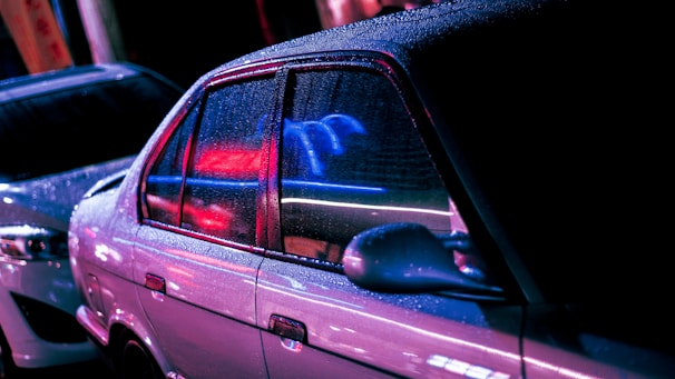 A nighttime city street scene with a modern car parked, lights reflecting tropical colors and neon effects.