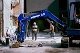 A construction site setting with a blue excavator prominently displayed. Two workers wearing casual attire are visible, with one standing in front of the machine and another partially visible in the seat of the excavator. Construction materials like wheelbarrows, pipes, and a partially demolished pavement are seen in the background.