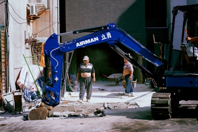 A construction site setting with a blue excavator prominently displayed. Two workers wearing casual attire are visible, with one standing in front of the machine and another partially visible in the seat of the excavator. Construction materials like wheelbarrows, pipes, and a partially demolished pavement are seen in the background.