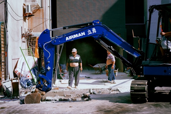 A construction site setting with a blue excavator prominently displayed. Two workers wearing casual attire are visible, with one standing in front of the machine and another partially visible in the seat of the excavator. Construction materials like wheelbarrows, pipes, and a partially demolished pavement are seen in the background.