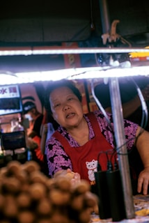 A woman in a red apron with a floral purple shirt is working at a food stall. She appears to be handing something over. A warm light illuminates her face and the surrounding area. There are other people slightly blurred in the background, and various food items are on display in the foreground.