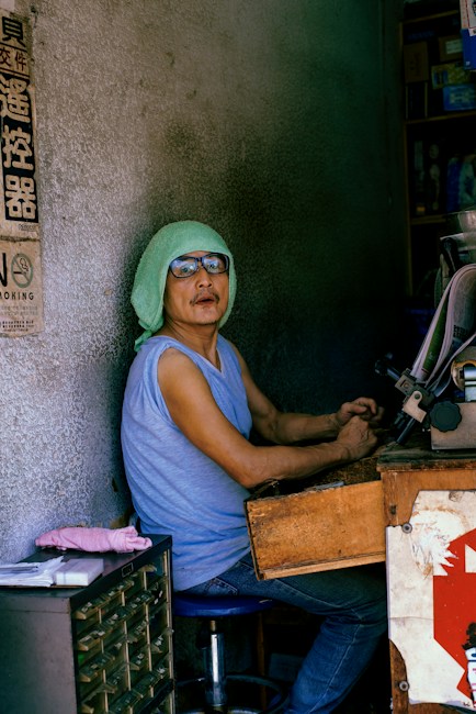 A person, likely an elderly man, is sitting inside a small, dimly-lit workspace. He is wearing glasses, a light blue sleeveless shirt, and a green towel on his head. The workspace looks cluttered with tools and equipment. There is a drawer unit to his left, with a pencil and some papers on top. The wall beside him has some signage, and the background includes some boxes and shelves.