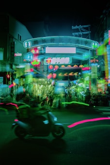 Neon-lit street scene at night with a prominent archway structure illuminated in green. Blurred motion of a scooter in the foreground and vibrant lights reflecting off buildings. Red lanterns are visible in the background, indicating possible festival or market events.
